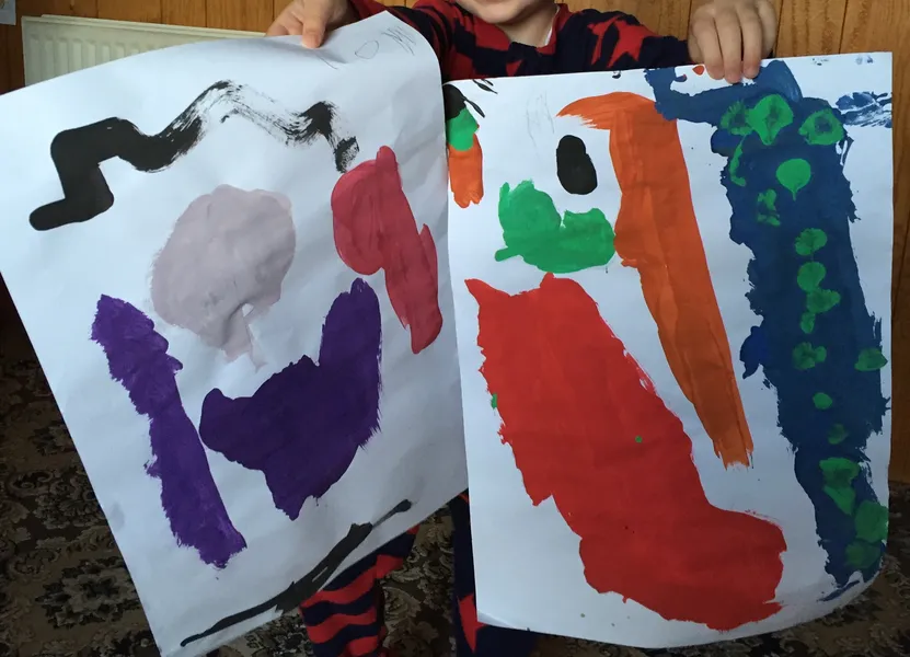 A child holding up two large abstract paintings in bold poster-paint colours